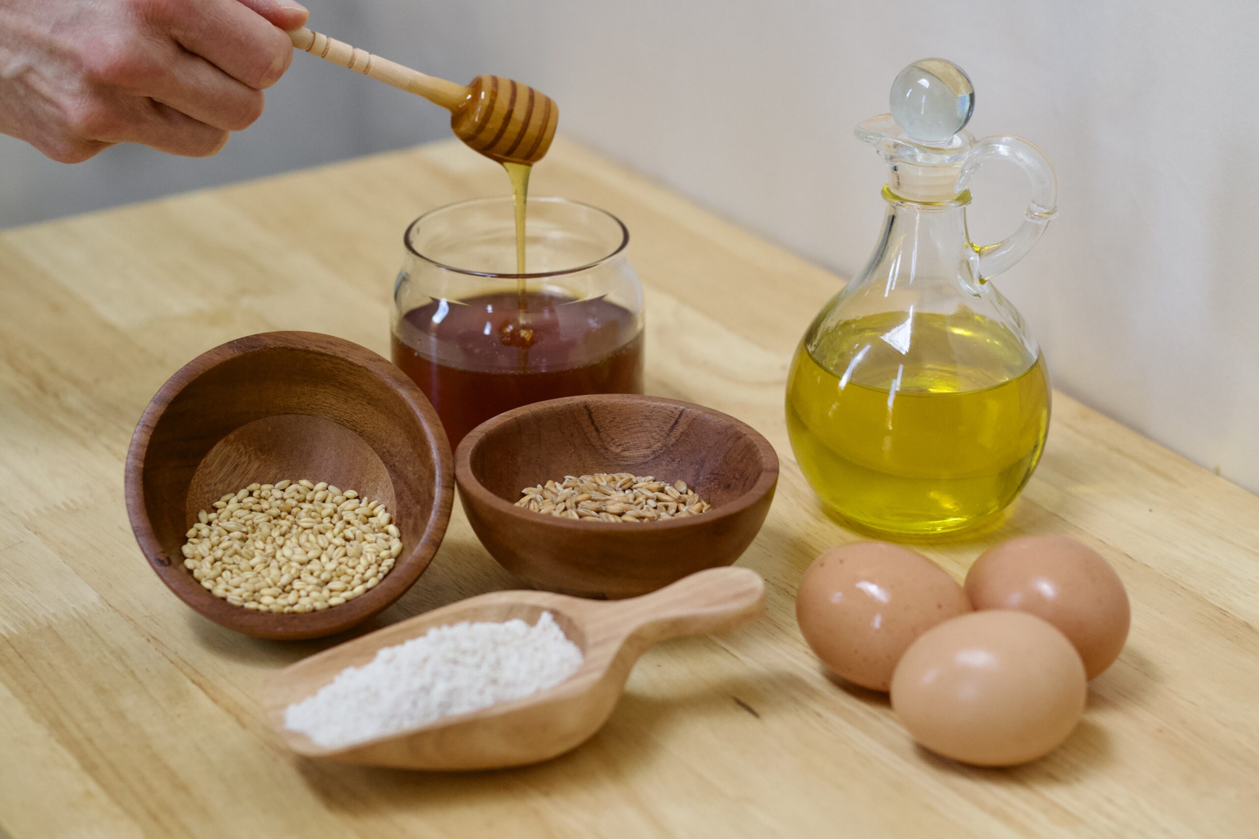 Photo of organic ingredients used to make whole grain hearth bread, including jar of honey with honey flowing from raised dipper into jar, small wooden bowl of hard red organic wheat berries, small wooden bowl of soft white wheat berries, small wooden scoop of freshly milled organic whole grain wheat flour, cruet of organic extra virgin olive oil, and three organic cage free brown eggs.