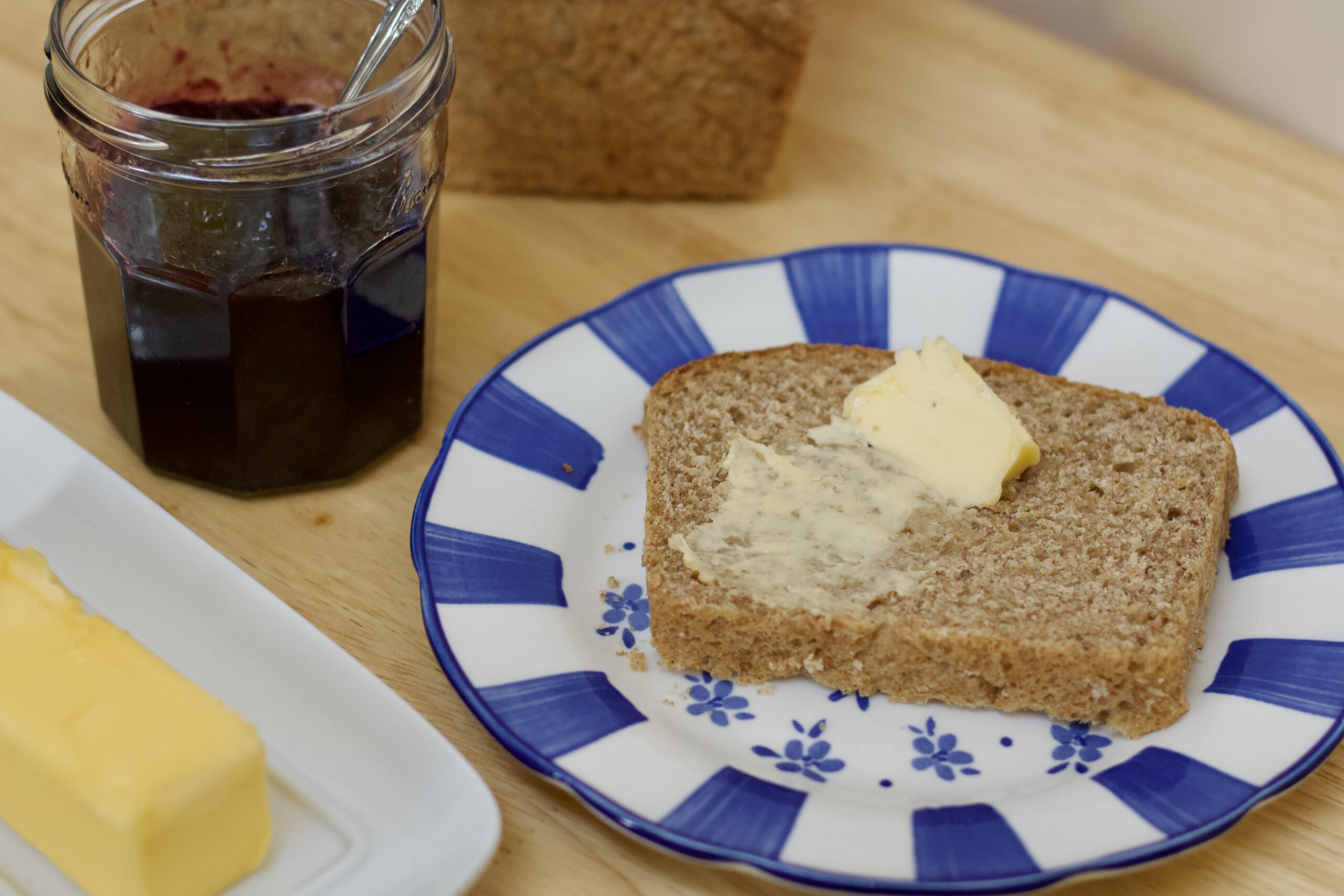 Photo of slice of whole grain hearth bread with butter on a blue and white patterned plate, white butter dish with gold butter, clear glass jar with blueberry preserves, and background image of whole grain hearth bread loaf.