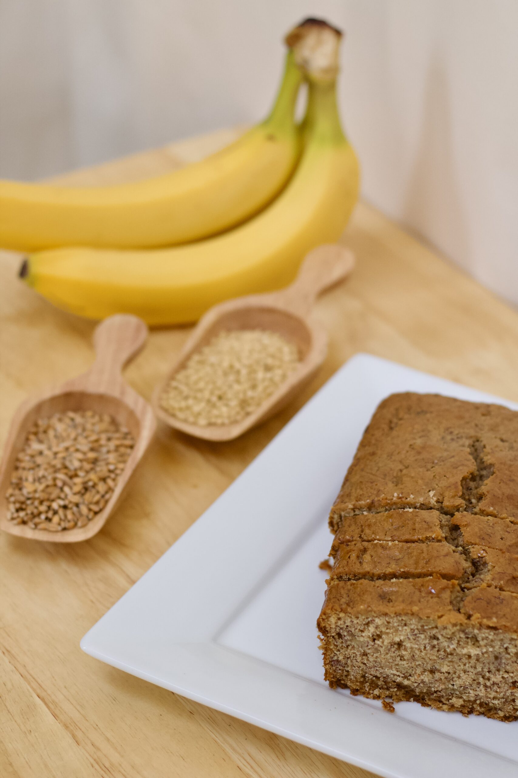 Photo of Brother Casper's whole grain banana bread with fresh organic bananas, small wooden scoop with organic spelt berries, small wooden scoop of organic soft white wheat berries, and banana bread with slices on shiny white serving plate.