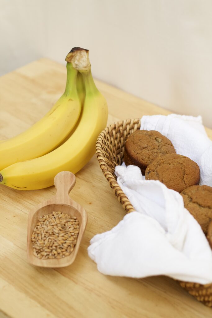 Photo of fresh bananas, wooden scoop of organic wheat berries, and cloth lined basket containing whole grain banana muffins made from organic ingredients.