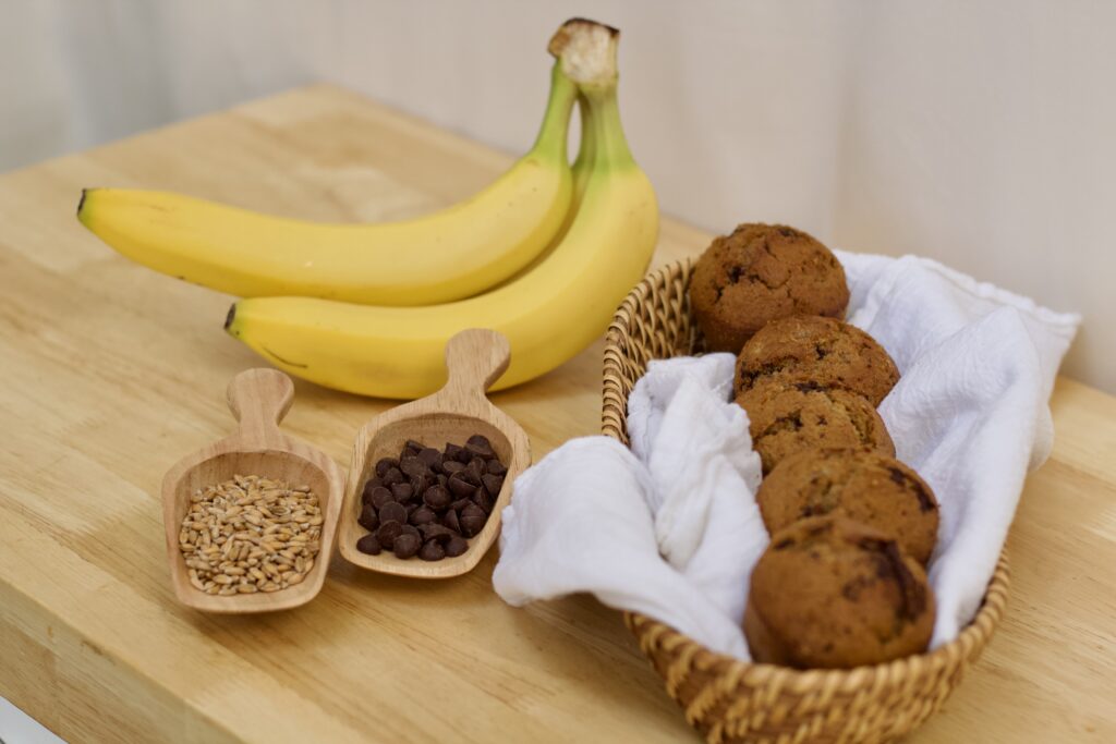 Photo of organic ingredients and finished whole grain chocolate banana feast day muffins, including ripe organic bananas, small wooden scoop of organic wheat berries, small wooden scoop of organic chocolate chips, and white cloth lined basked of finished muffins.