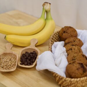 Photo of organic ingredients and finished whole grain chocolate banana feast day muffins, including ripe organic bananas, small wooden scoop of organic wheat berries, small wooden scoop of organic chocolate chips, and white cloth lined basked of finished muffins.