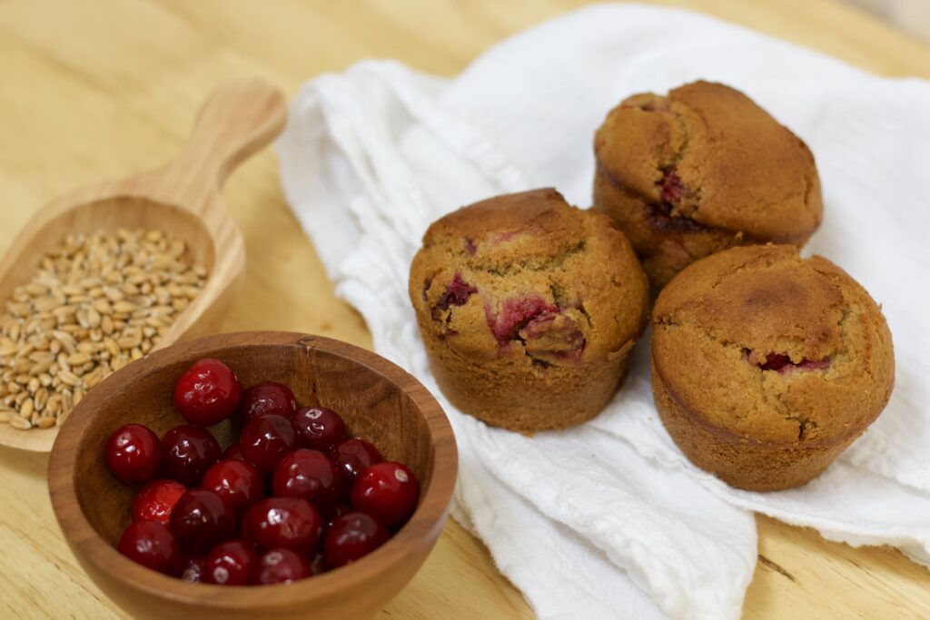 Photo of small wooden scoop containing organic wheat berries, small wooden bowl of glistening organic cranberries, and white cloth under cranberry's glory whole grain muffins made with organic ingredients.
