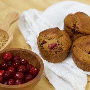 Photo of small wooden scoop containing organic wheat berries, small wooden bowl of glistening organic cranberries, and white cloth under cranberry's glory whole grain muffins made with organic ingredients.