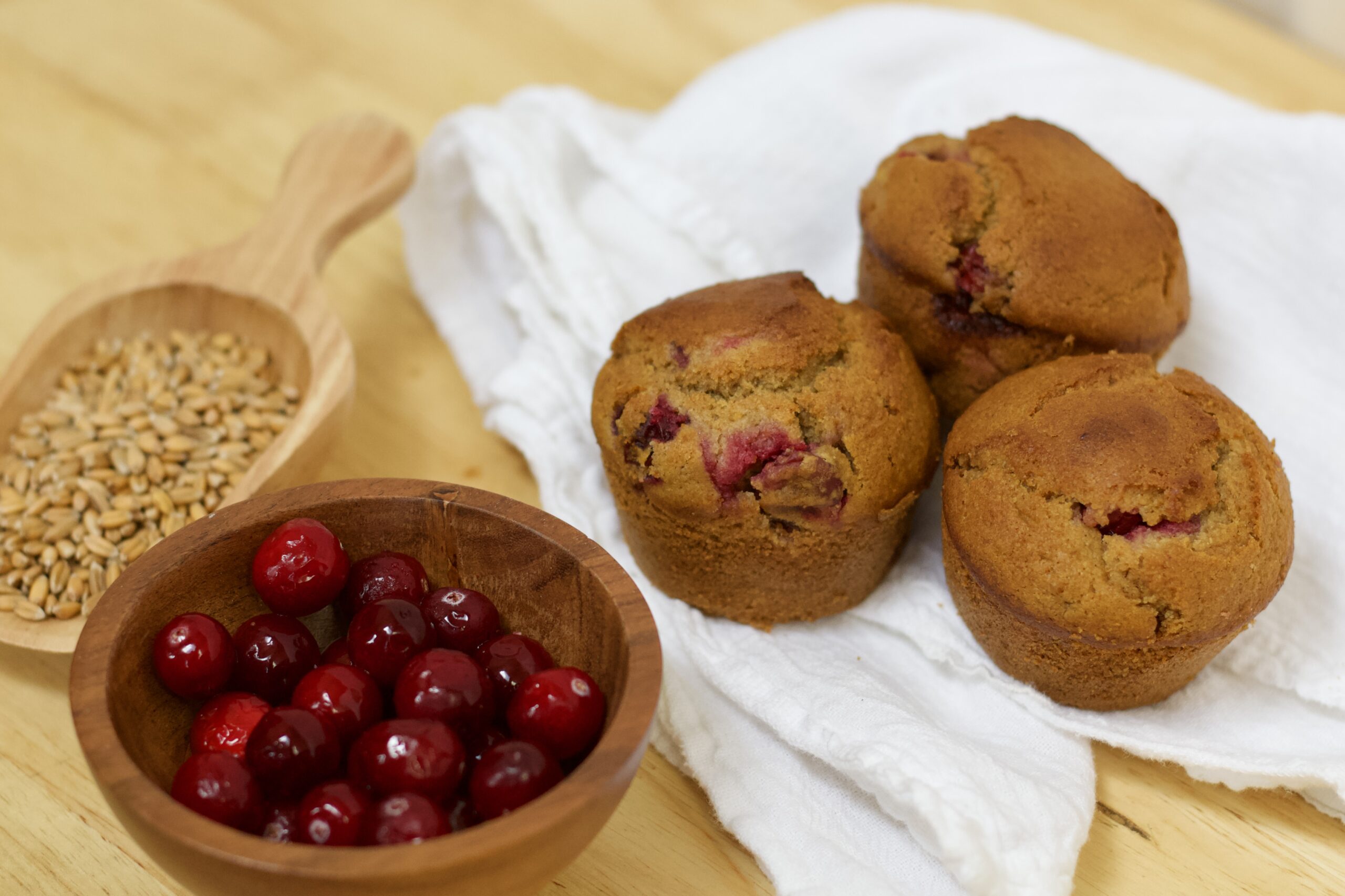 Photo of small wooden scoop containing organic wheat berries, small wooden bowl of glistening organic cranberries, and white cloth under cranberry's glory whole grain muffins made with organic ingredients.