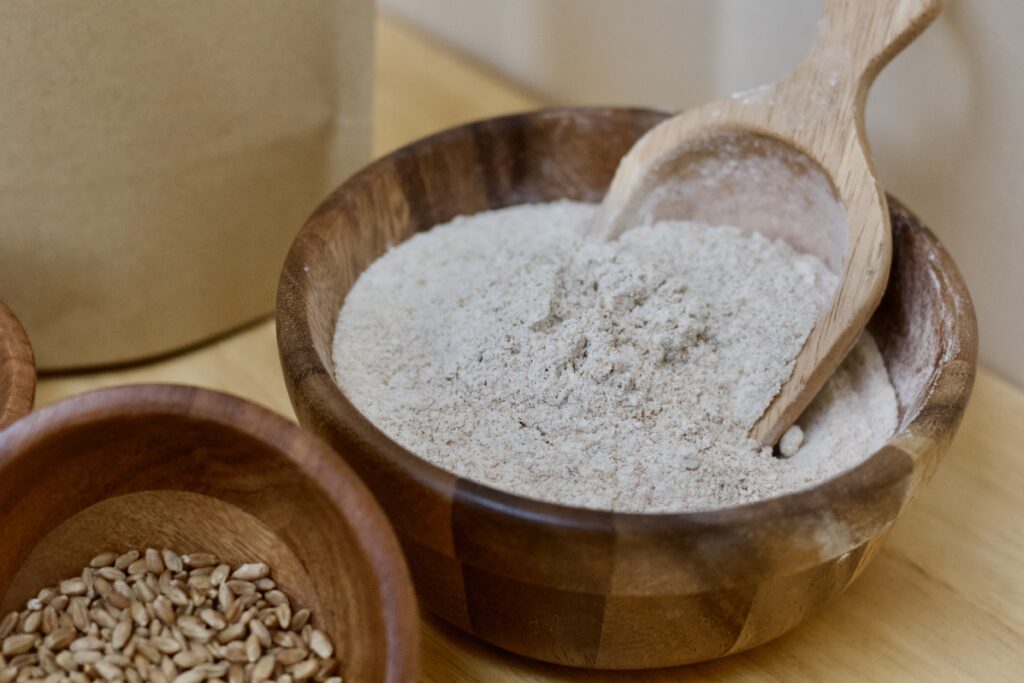 Photo focusing of medium wooden bowl with small wooden scoop that contains fresh milled flour blend, small section of small wooden bowl containing organic red wheat berries, and section of kraft flour bag.