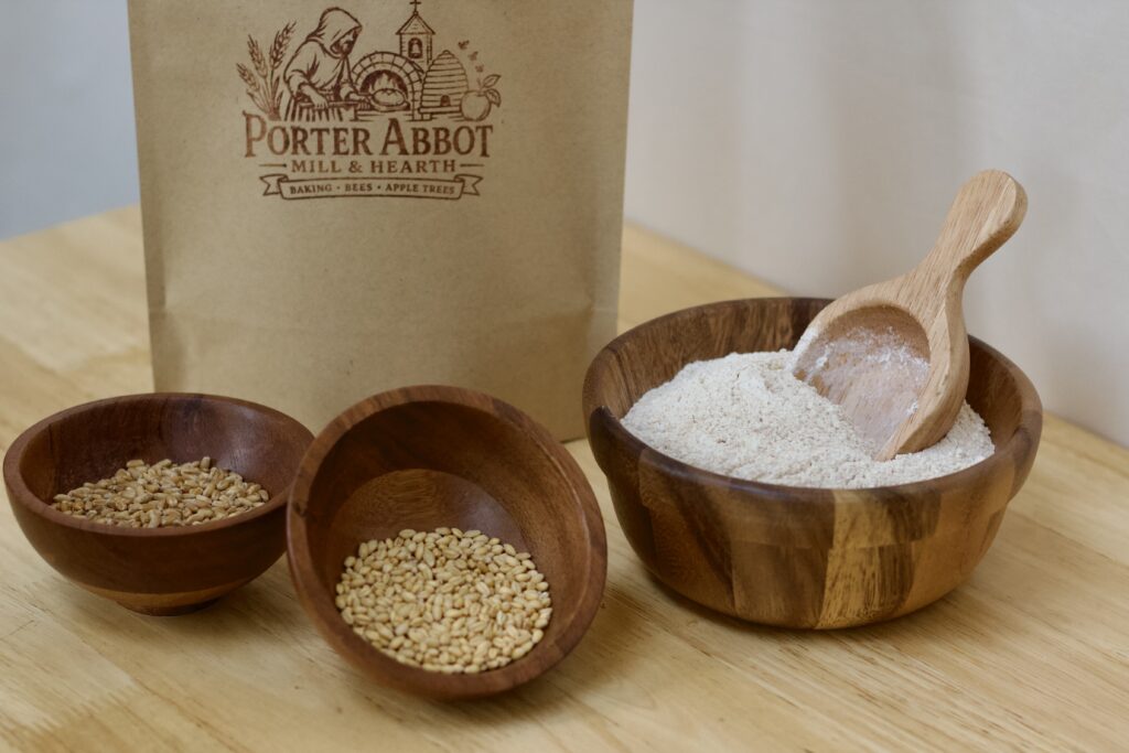 Photo of freshly milled whole grain white wheat flour blend, including sealable kraft flour bag, small wooden bowl of organic soft white wheat berries, small wooden bowl of organic hard white wheat berries, medium wooden bowl of freshly milled whole grain white wheat flour blend and small wooden scoop.