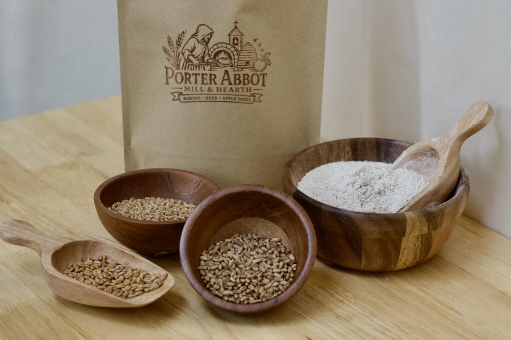 Photo of freshly milled organic whole grain red wheat flour blend, including sealable kraft flour bag, small wooden scoop of soft white wheat berries, small wooden bowl of heirloom organic red wheat berries, small wooden bowl of organic red wheat berries, and medium wooden bowl of whole grain flour with small wooden scoop.