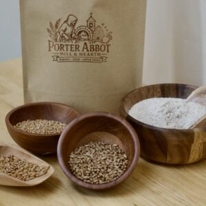 Photo of freshly milled organic whole grain red wheat flour blend, including sealable kraft flour bag, small wooden scoop of soft white wheat berries, small wooden bowl of heirloom organic red wheat berries, small wooden bowl of organic red wheat berries, and medium wooden bowl of whole grain flour with small wooden scoop.